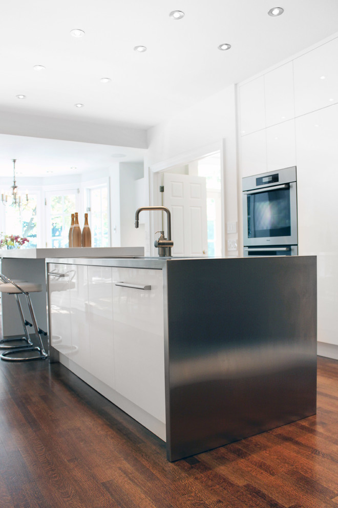 This is an example of a contemporary kitchen in Toronto with an integrated sink, flat-panel cabinets, white cabinets, stainless steel worktops, white splashback, glass sheet splashback, integrated appliances and medium hardwood flooring.