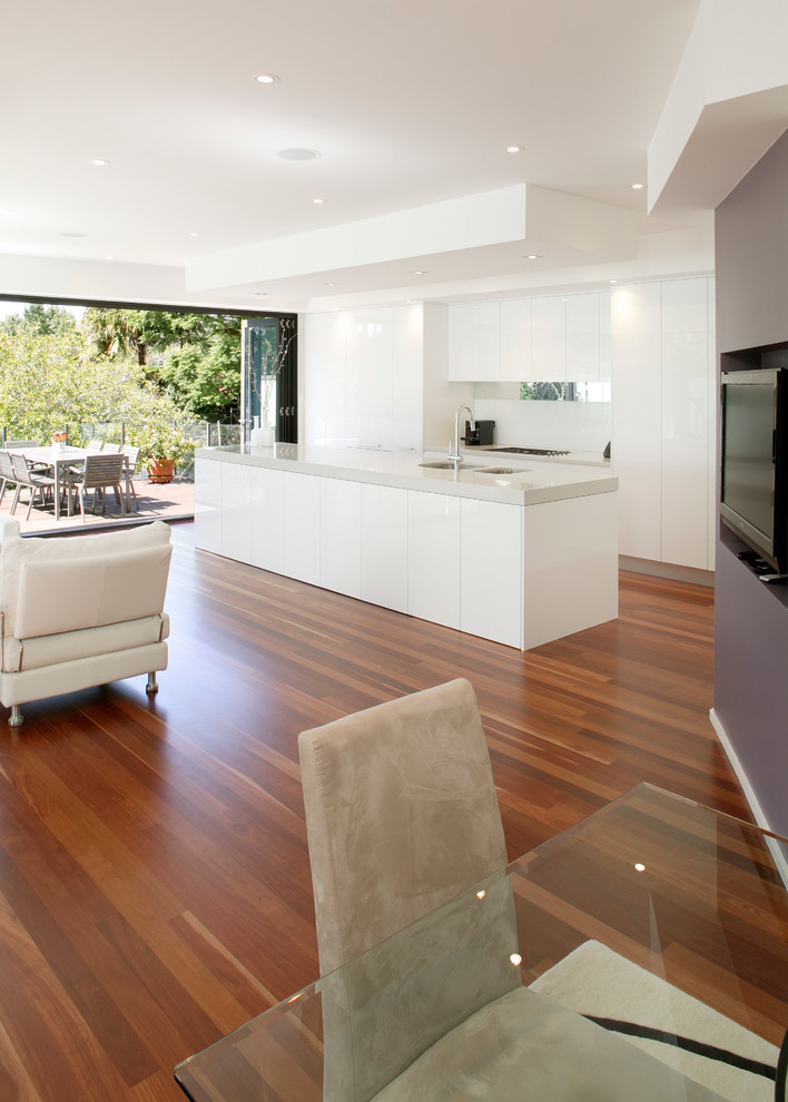 Example of a minimalist galley medium tone wood floor eat-in kitchen design in Sydney with a drop-in sink, flat-panel cabinets, white cabinets, quartz countertops, white backsplash, glass tile backsplash, stainless steel appliances and an island