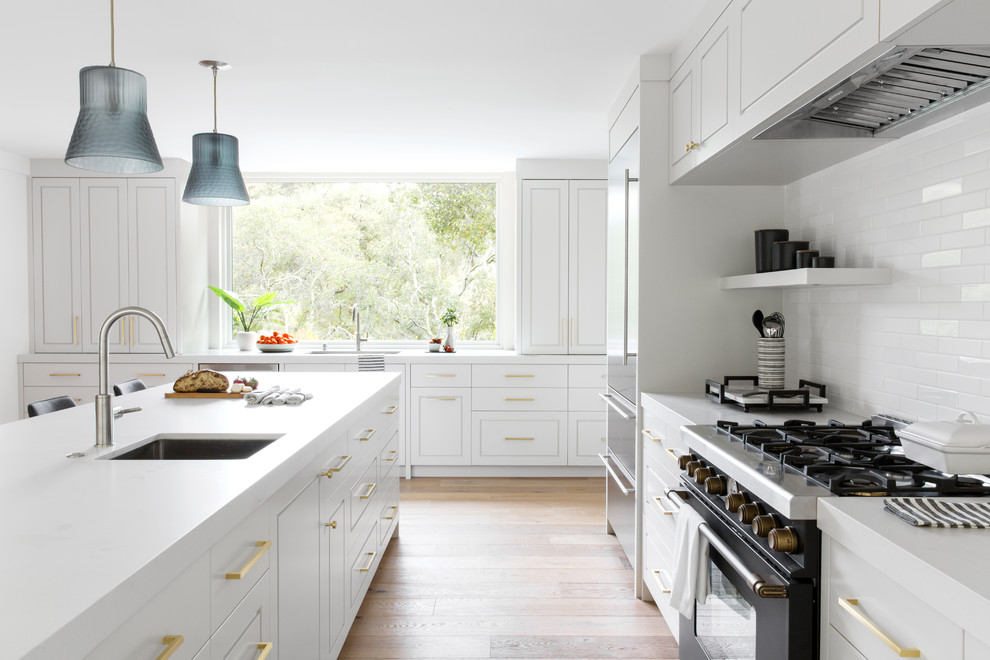This is an example of a large traditional l-shaped kitchen in San Francisco with a single-bowl sink, engineered stone countertops, white splashback, ceramic splashback, black appliances, light hardwood flooring, an island, grey worktops, raised-panel cabinets and beige floors.