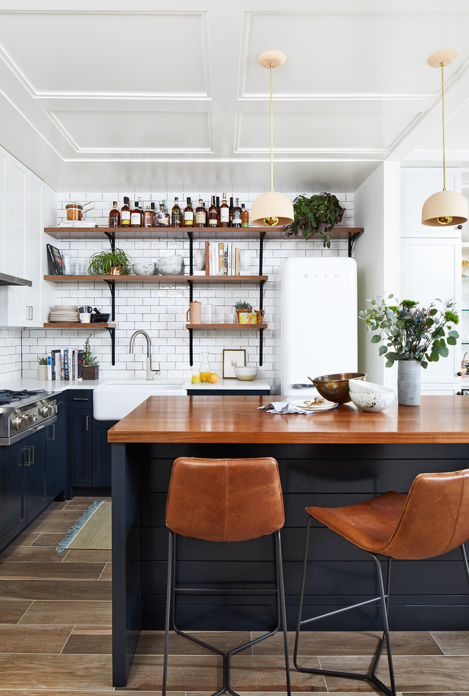 Small trendy l-shaped porcelain tile and brown floor open concept kitchen photo in DC Metro with a farmhouse sink, shaker cabinets, blue cabinets, wood countertops, white backsplash, ceramic backsplash, an island, white appliances and brown countertops