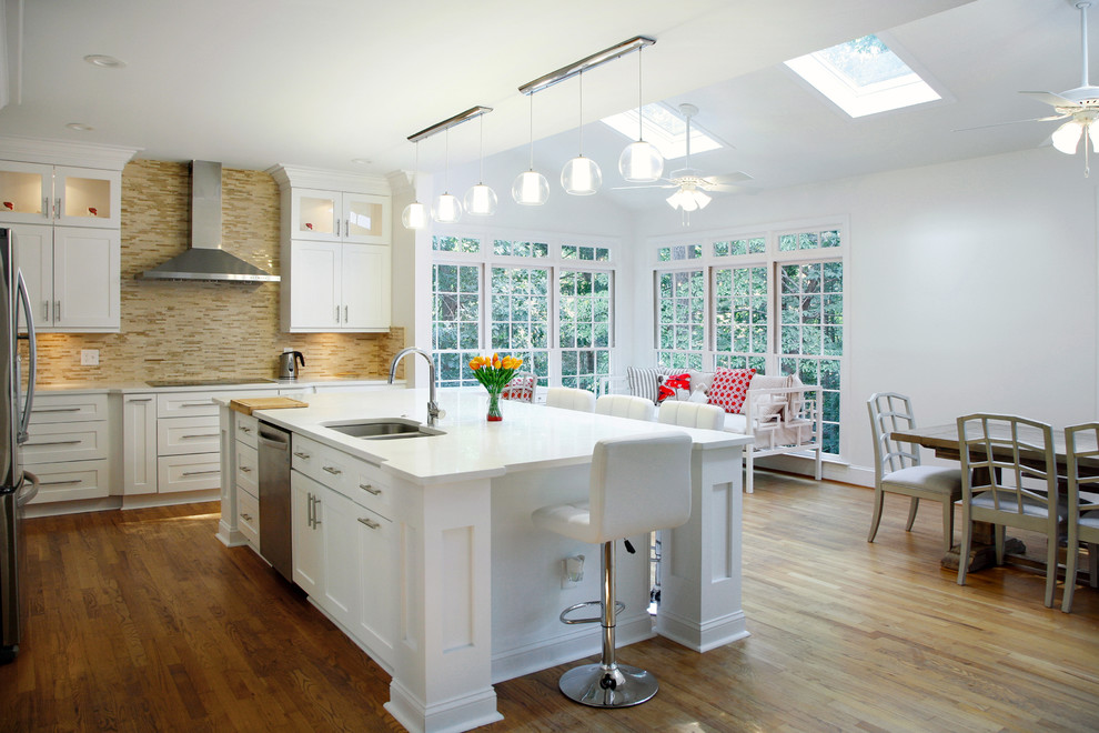 This is an example of a large traditional l-shaped kitchen/diner in Atlanta with a submerged sink, shaker cabinets, beige splashback, stone tiled splashback, stainless steel appliances, quartz worktops, an island, white cabinets, medium hardwood flooring, brown floors and white worktops.