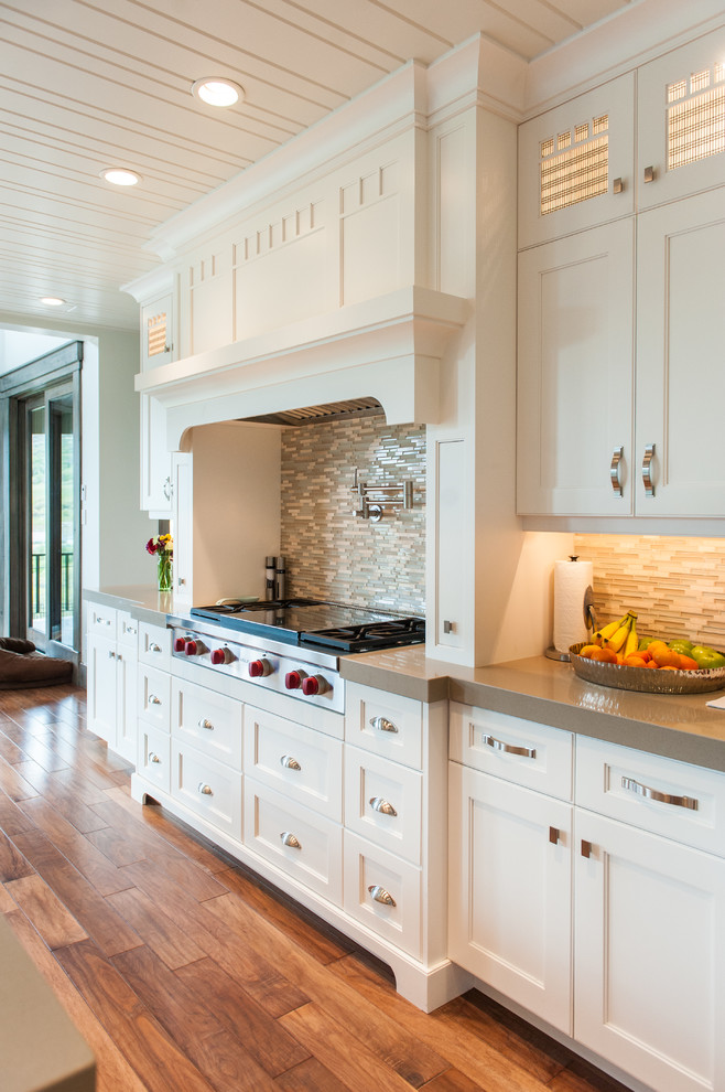 Photo of a large classic l-shaped open plan kitchen in Salt Lake City with white cabinets, a belfast sink, shaker cabinets, composite countertops, multi-coloured splashback, glass tiled splashback, integrated appliances, medium hardwood flooring, an island and brown floors.