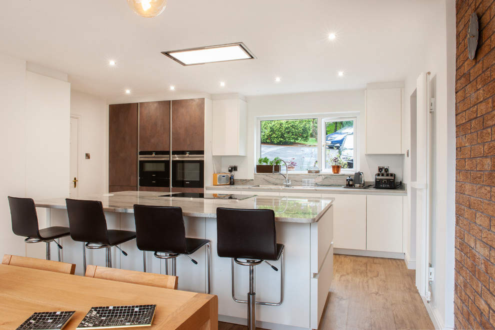 This is an example of a large contemporary open plan kitchen in Manchester with flat-panel cabinets, white cabinets, quartz worktops, black appliances, vinyl flooring and an island.