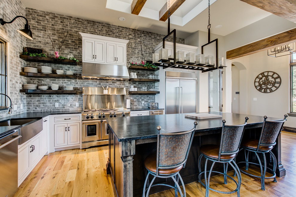 This is an example of a medium sized industrial l-shaped open plan kitchen in Dallas with a belfast sink, recessed-panel cabinets, white cabinets, granite worktops, grey splashback, metro tiled splashback, stainless steel appliances, light hardwood flooring, an island, brown floors and black worktops.