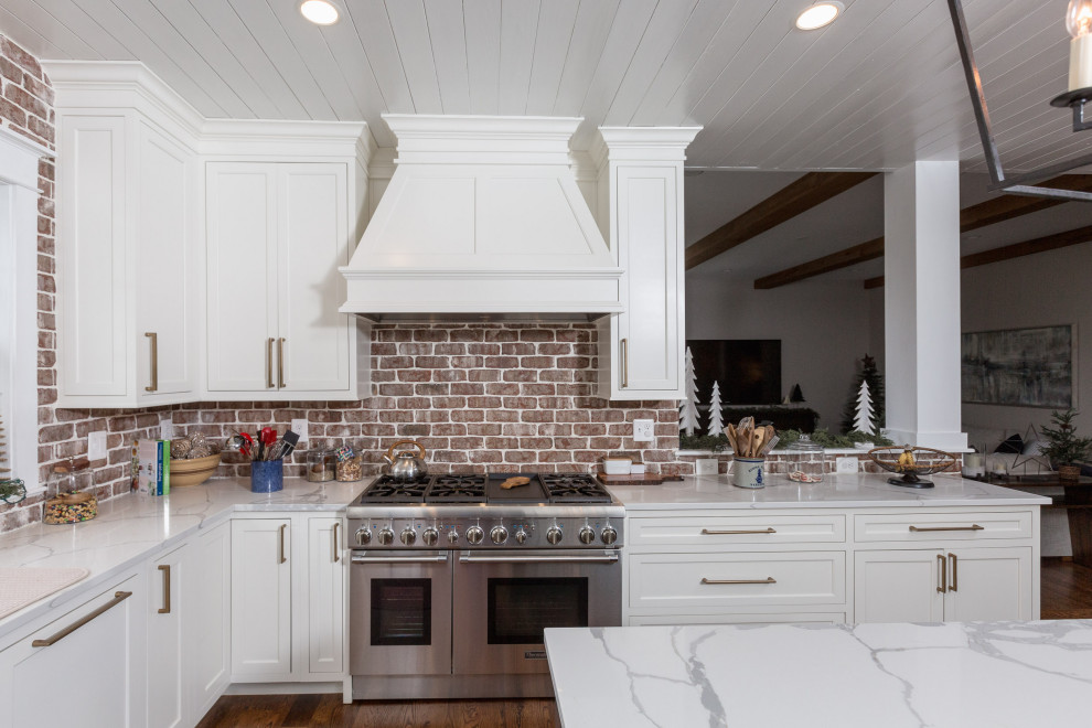 Inredning av ett klassiskt stort vit vitt kök, med en rustik diskho, skåp i shakerstil, vita skåp, bänkskiva i kvarts, rött stänkskydd, stänkskydd i tegel, rostfria vitvaror, mörkt trägolv, en köksö och brunt golv