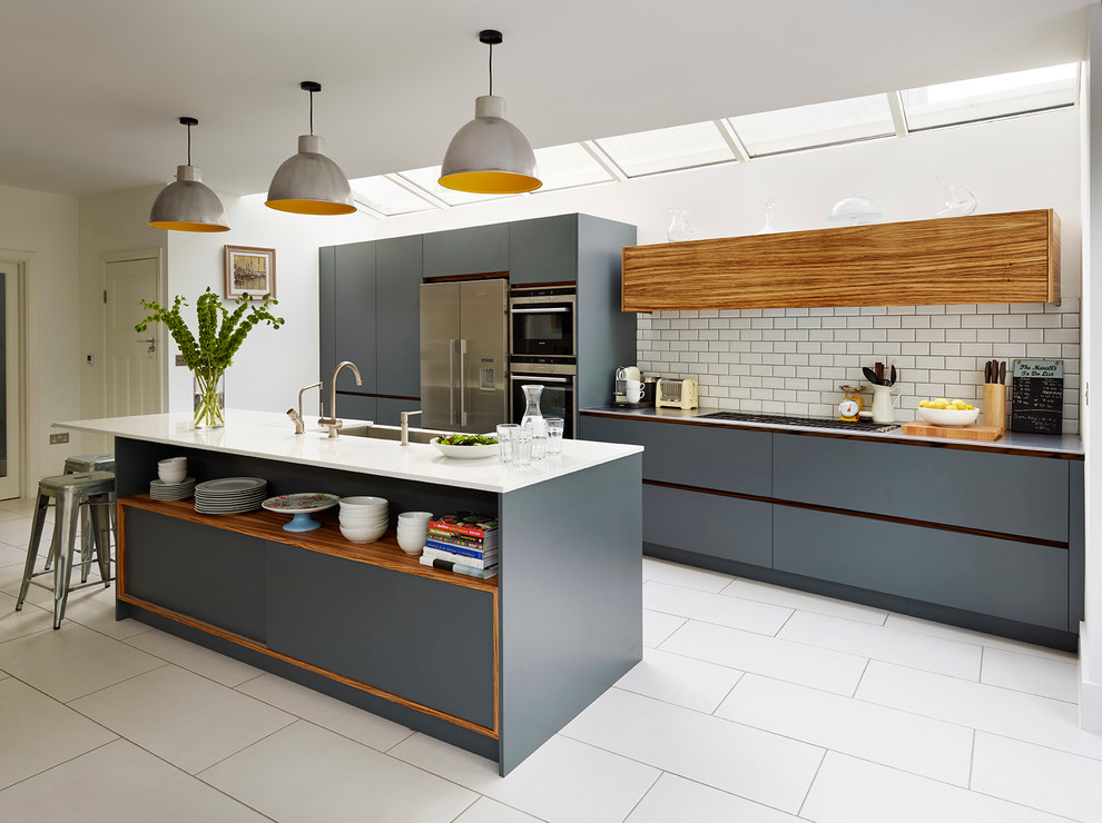 Example of a large trendy open concept kitchen design in London with an undermount sink, flat-panel cabinets, gray cabinets, solid surface countertops, white backsplash, ceramic backsplash, stainless steel appliances and an island