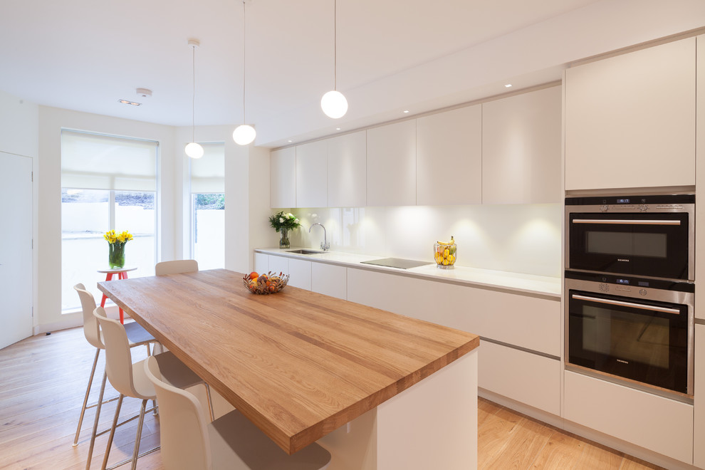 Large contemporary open plan kitchen in London with a submerged sink, flat-panel cabinets, white cabinets, composite countertops, white splashback, glass sheet splashback and an island.