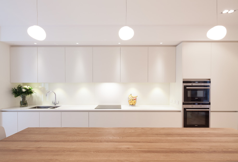 Design ideas for a large contemporary open plan kitchen in London with a submerged sink, flat-panel cabinets, white cabinets, composite countertops, white splashback, glass sheet splashback and an island.