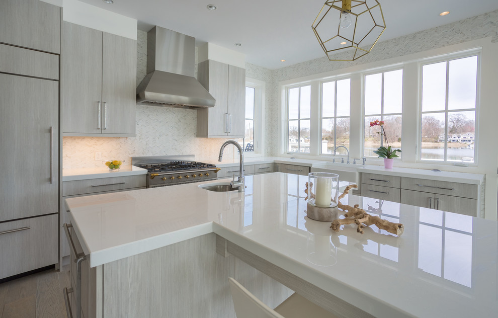 This is an example of a large contemporary l-shaped open plan kitchen in New York with a belfast sink, flat-panel cabinets, grey cabinets, glass worktops, grey splashback, stone tiled splashback, stainless steel appliances, medium hardwood flooring and an island.