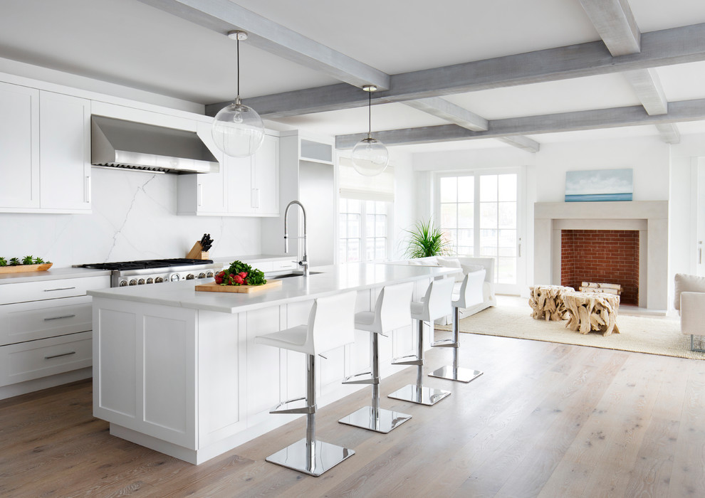 Cette photo montre une cuisine américaine parallèle bord de mer de taille moyenne avec un placard à porte shaker, des portes de placard blanches, une crédence blanche, un électroménager en acier inoxydable, un sol en bois brun, îlot, un sol marron et une crédence en marbre.