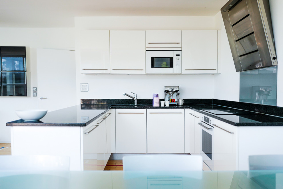 Inspiration for a small contemporary u-shaped open plan kitchen in London with a built-in sink, flat-panel cabinets, white cabinets, granite worktops, grey splashback, glass sheet splashback, white appliances, light hardwood flooring, no island, multi-coloured floors, black worktops and a drop ceiling.