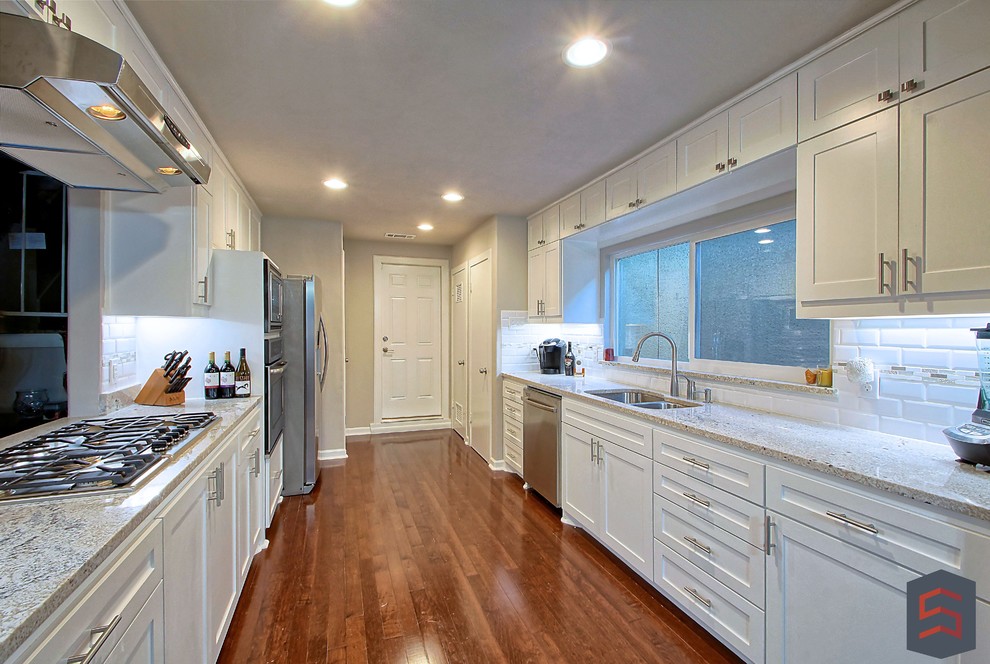 Example of a mid-sized transitional galley dark wood floor enclosed kitchen design in Dallas with white cabinets, granite countertops, white backsplash, stainless steel appliances, a double-bowl sink, shaker cabinets and subway tile backsplash