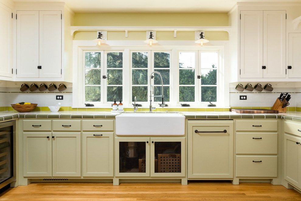 This is an example of a classic u-shaped enclosed kitchen in Los Angeles with a belfast sink, shaker cabinets, green cabinets, tile countertops, white splashback, metro tiled splashback, stainless steel appliances and medium hardwood flooring.