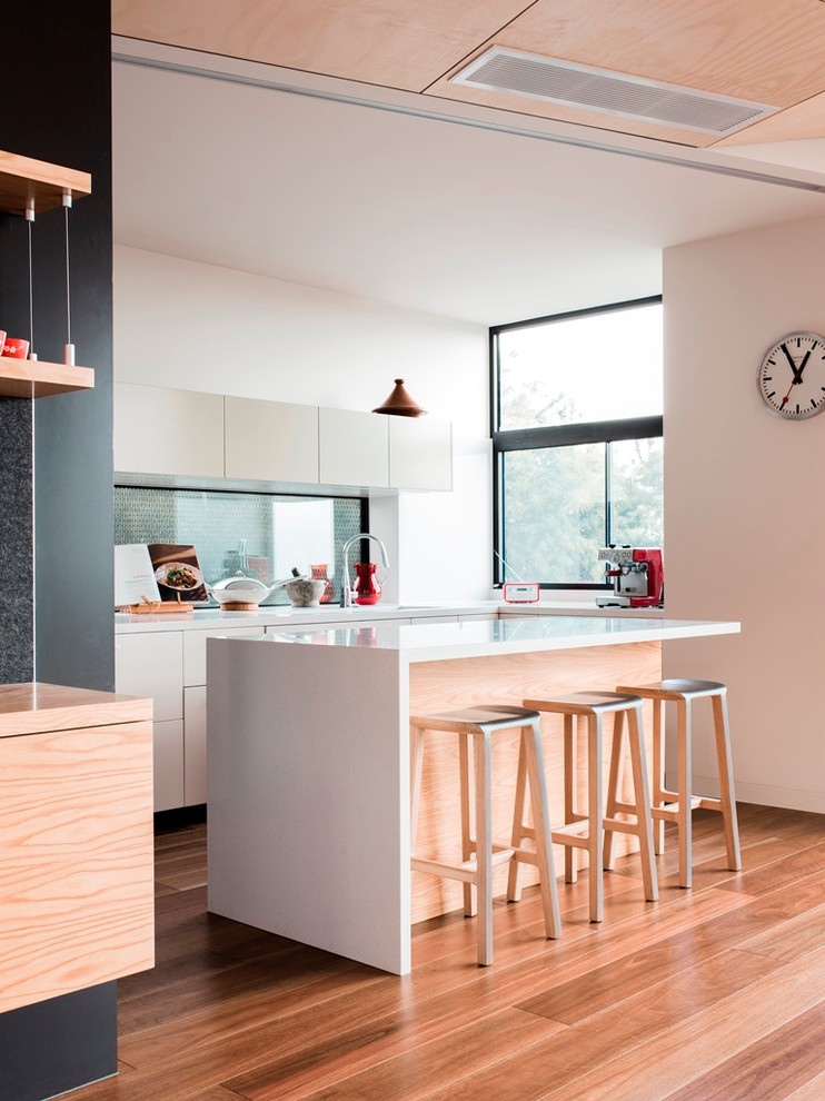 Idée de décoration pour une cuisine design en L de taille moyenne avec un évier encastré, un placard à porte plane, des portes de placard blanches, un plan de travail en quartz modifié, une crédence en feuille de verre, un sol en bois brun et îlot.