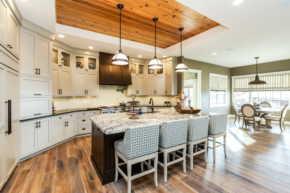 Cette image montre une cuisine américaine traditionnelle avec un placard à porte shaker, des portes de placard beiges, une crédence blanche, un sol en bois brun et îlot.