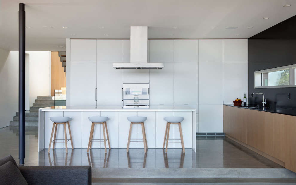 На фото: угловая кухня-гостиная в стиле модернизм с врезной мойкой, плоскими фасадами, белыми фасадами, черным фартуком и бетонным полом