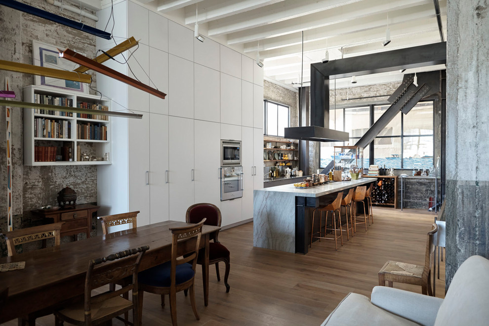 Industrial galley kitchen/diner in San Francisco with flat-panel cabinets, white cabinets, stainless steel appliances, medium hardwood flooring, an island and brown floors.