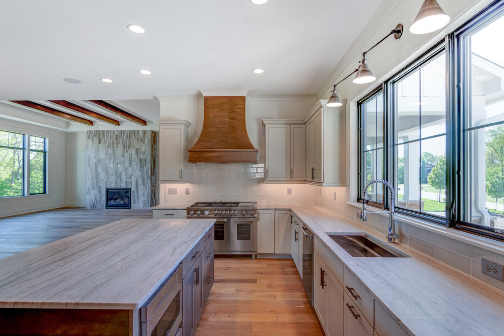 Photo of a rural l-shaped kitchen/diner in Indianapolis with a belfast sink, shaker cabinets, grey cabinets, quartz worktops, white splashback, ceramic splashback, stainless steel appliances, light hardwood flooring, an island, brown floors and grey worktops.