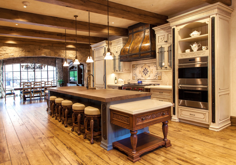 Inspiration for a french country galley medium tone wood floor, brown floor and exposed beam eat-in kitchen remodel in New York with a farmhouse sink, raised-panel cabinets, distressed cabinets, wood countertops, multicolored backsplash, paneled appliances, an island and brown countertops