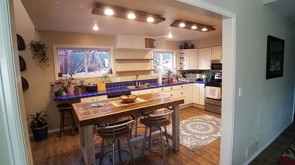 Large rustic l-shaped kitchen/diner in Denver with a double-bowl sink, raised-panel cabinets, white cabinets, tile countertops, blue splashback, ceramic splashback, stainless steel appliances, light hardwood flooring, an island and brown floors.