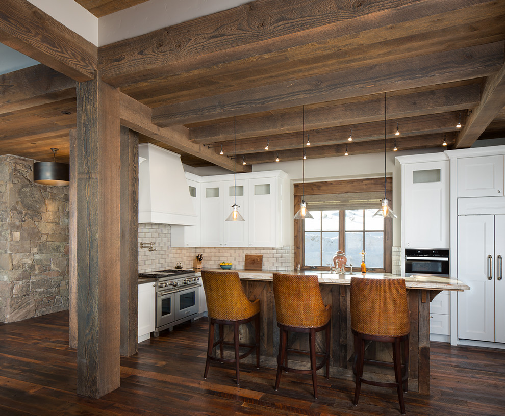Kitchen - rustic l-shaped dark wood floor and brown floor kitchen idea in Salt Lake City with recessed-panel cabinets, white cabinets, beige backsplash, subway tile backsplash, paneled appliances and an island
