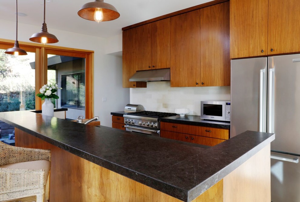 Modern inredning av ett mellanstort kök, med släta luckor, skåp i mellenmörkt trä, granitbänkskiva, vitt stänkskydd, stänkskydd i porslinskakel, rostfria vitvaror och en köksö