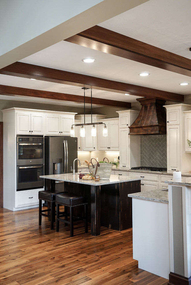 Photo of a medium sized country l-shaped kitchen/diner in Other with a submerged sink, raised-panel cabinets, white cabinets, quartz worktops, grey splashback, metro tiled splashback, stainless steel appliances, an island, brown floors and medium hardwood flooring.