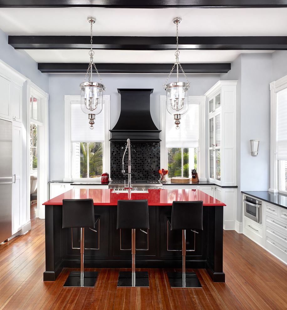 Geschlossene Klassische Küche in L-Form mit Landhausspüle, weißen Schränken, Quarzwerkstein-Arbeitsplatte, Küchenrückwand in Schwarz, Küchengeräten aus Edelstahl, braunem Holzboden, Kücheninsel, Glasfronten und roter Arbeitsplatte in Charleston