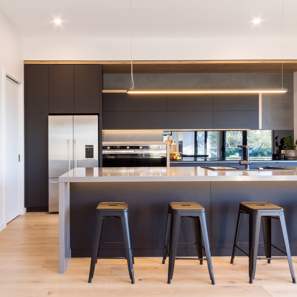 Inspiration for a medium sized modern galley kitchen/diner in Melbourne with a built-in sink, flat-panel cabinets, black cabinets, engineered stone countertops, black splashback, mirror splashback, stainless steel appliances, light hardwood flooring, an island, beige floors and grey worktops.