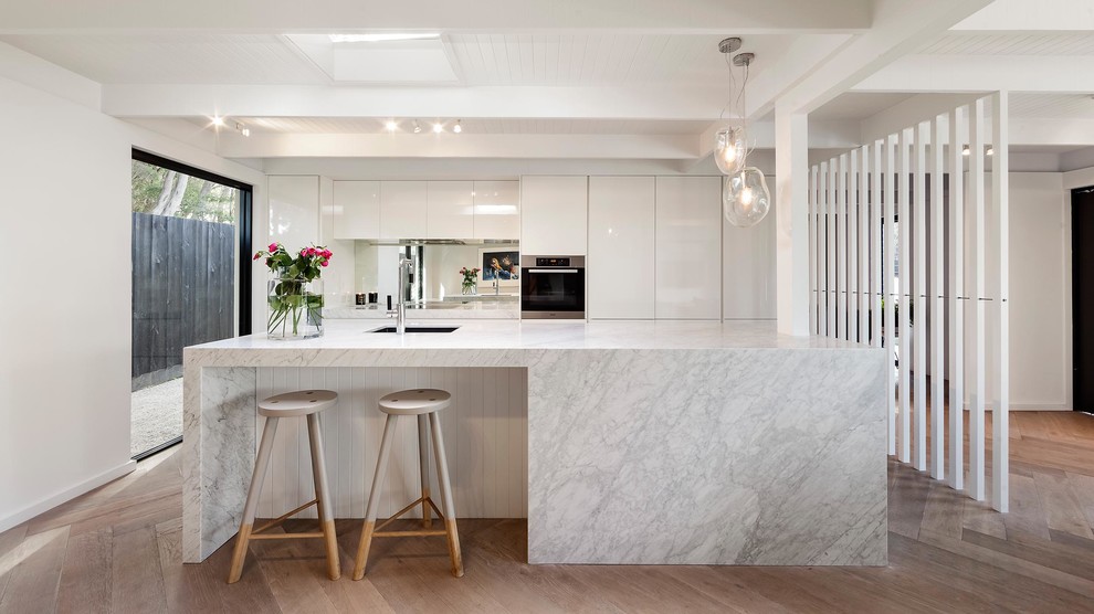 Open concept kitchen - contemporary galley light wood floor open concept kitchen idea in Melbourne with an undermount sink, flat-panel cabinets, white cabinets, metallic backsplash, mirror backsplash, stainless steel appliances and an island