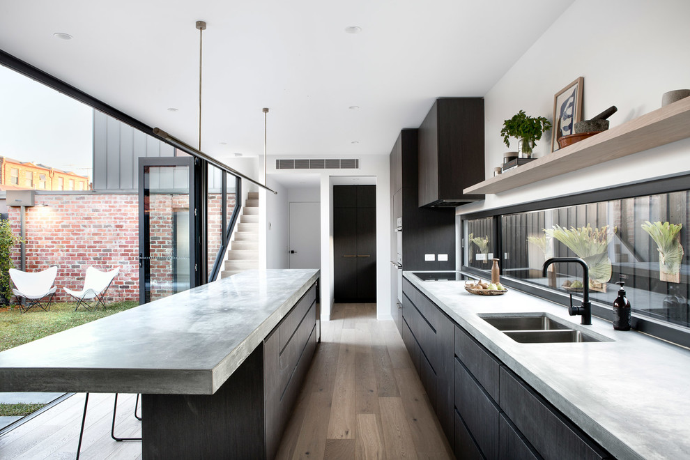 This is an example of a modern galley kitchen in Melbourne with a submerged sink, flat-panel cabinets, dark wood cabinets, concrete worktops, window splashback, integrated appliances, medium hardwood flooring, an island, beige floors and grey worktops.