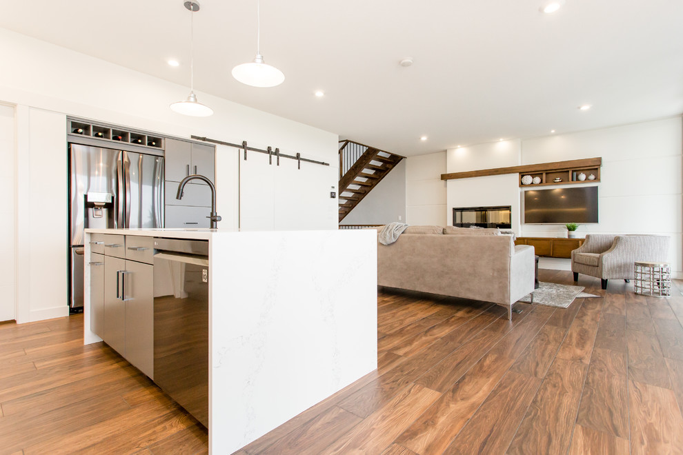 Photo of a medium sized contemporary l-shaped kitchen/diner in Calgary with a submerged sink, flat-panel cabinets, grey cabinets, engineered stone countertops, white splashback, stone slab splashback, stainless steel appliances, vinyl flooring, an island, brown floors and white worktops.