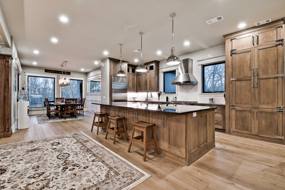 Inspiration for a huge modern light wood floor kitchen remodel in Other with a farmhouse sink, recessed-panel cabinets, brown cabinets, granite countertops, white backsplash, porcelain backsplash, stainless steel appliances, an island and black countertops