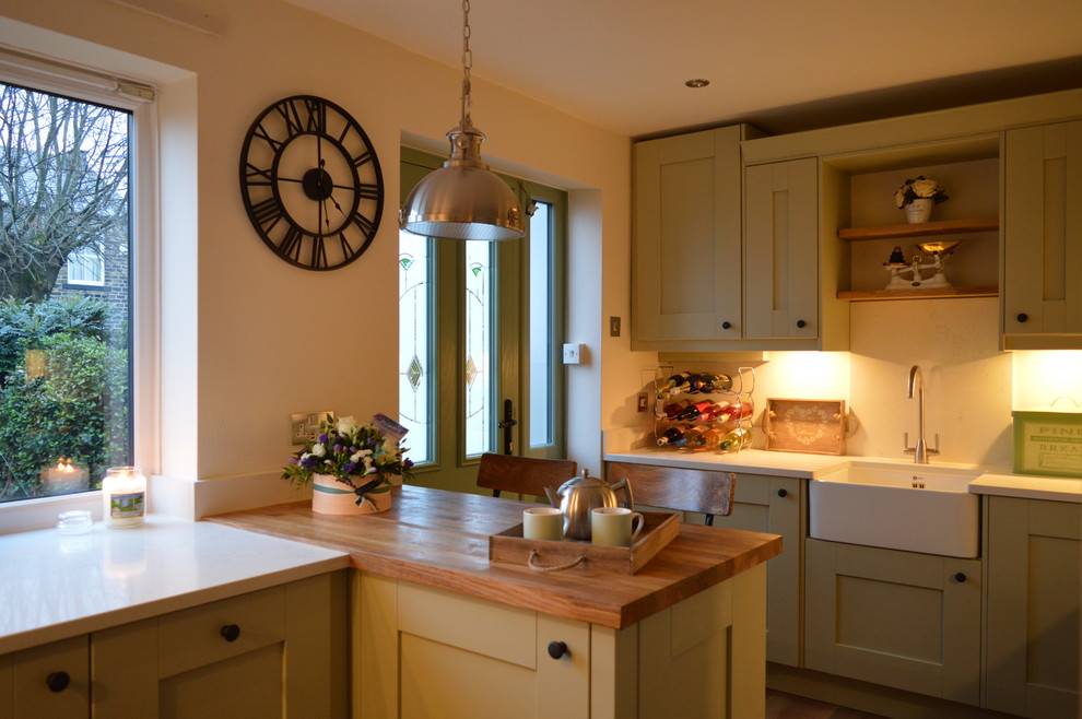 This is an example of a small romantic u-shaped enclosed kitchen in Other with a belfast sink, shaker cabinets, green cabinets, quartz worktops, white splashback, stone slab splashback, integrated appliances, laminate floors, a breakfast bar, brown floors and white worktops.