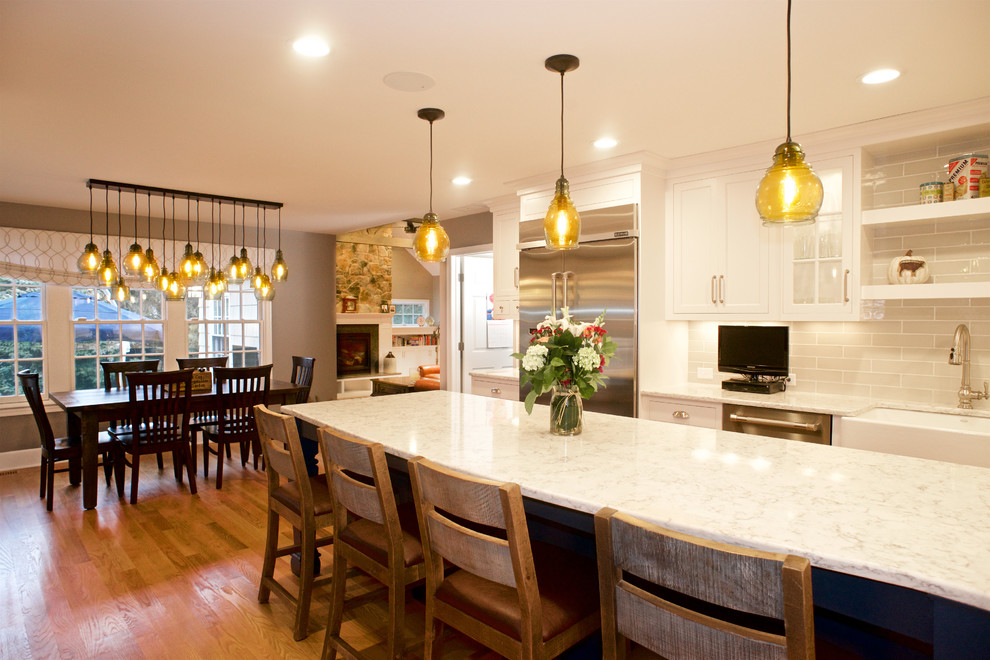Mid-sized beach style l-shaped medium tone wood floor eat-in kitchen photo in New York with a farmhouse sink, shaker cabinets, blue cabinets, quartz countertops, white backsplash, ceramic backsplash, stainless steel appliances, an island and white countertops