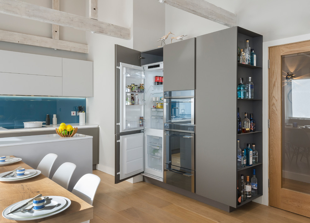 This is an example of a medium sized modern open plan kitchen in Devon with an integrated sink, flat-panel cabinets, composite countertops, blue splashback, glass sheet splashback, stainless steel appliances, light hardwood flooring and an island.