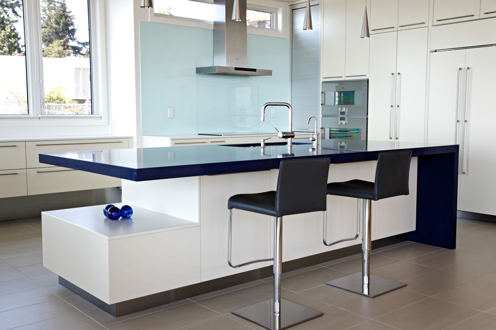 На фото: кухня в стиле модернизм с техникой под мебельный фасад, плоскими фасадами, белыми фасадами, синим фартуком, фартуком из стекла и синей столешницей