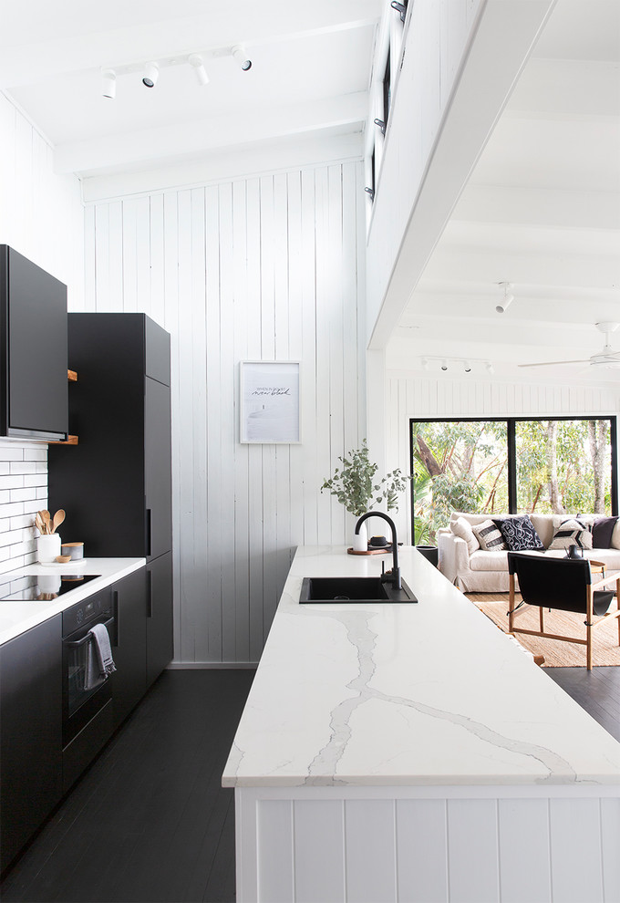 На фото: параллельная кухня-гостиная в морском стиле с накладной мойкой, плоскими фасадами, черными фасадами, белым фартуком, черной техникой, деревянным полом, полуостровом, черным полом и белой столешницей с