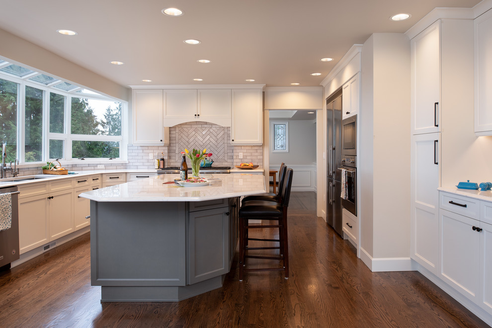 Inspiration pour une grande cuisine américaine traditionnelle en U avec un évier 3 bacs, un placard à porte shaker, des portes de placard grises, un plan de travail en quartz modifié, une crédence beige, une crédence en céramique, un électroménager en acier inoxydable, parquet foncé, îlot, un sol marron et un plan de travail blanc.