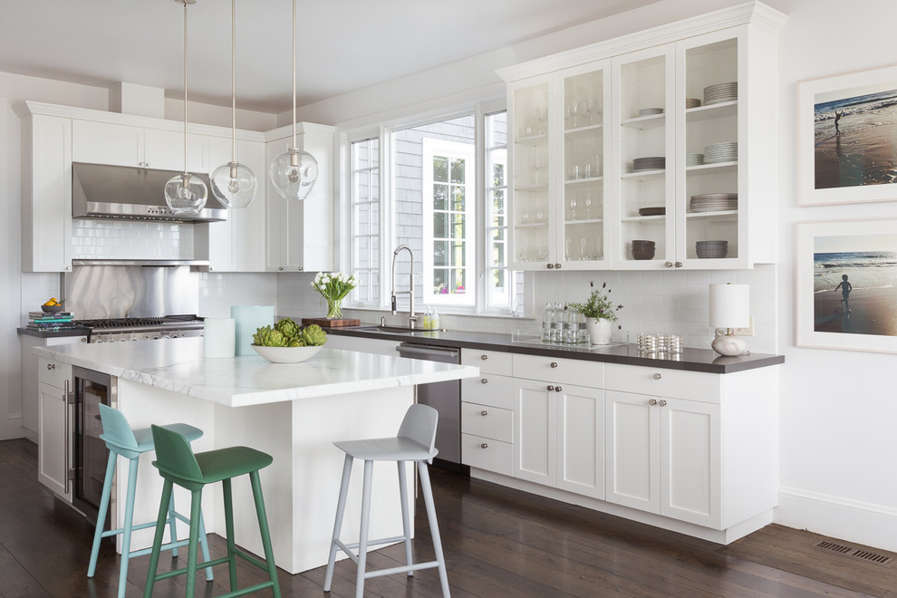 Exempel på ett klassiskt l-kök, med en undermonterad diskho, luckor med glaspanel, vita skåp, vitt stänkskydd, stänkskydd i tunnelbanekakel, rostfria vitvaror, mörkt trägolv och en köksö