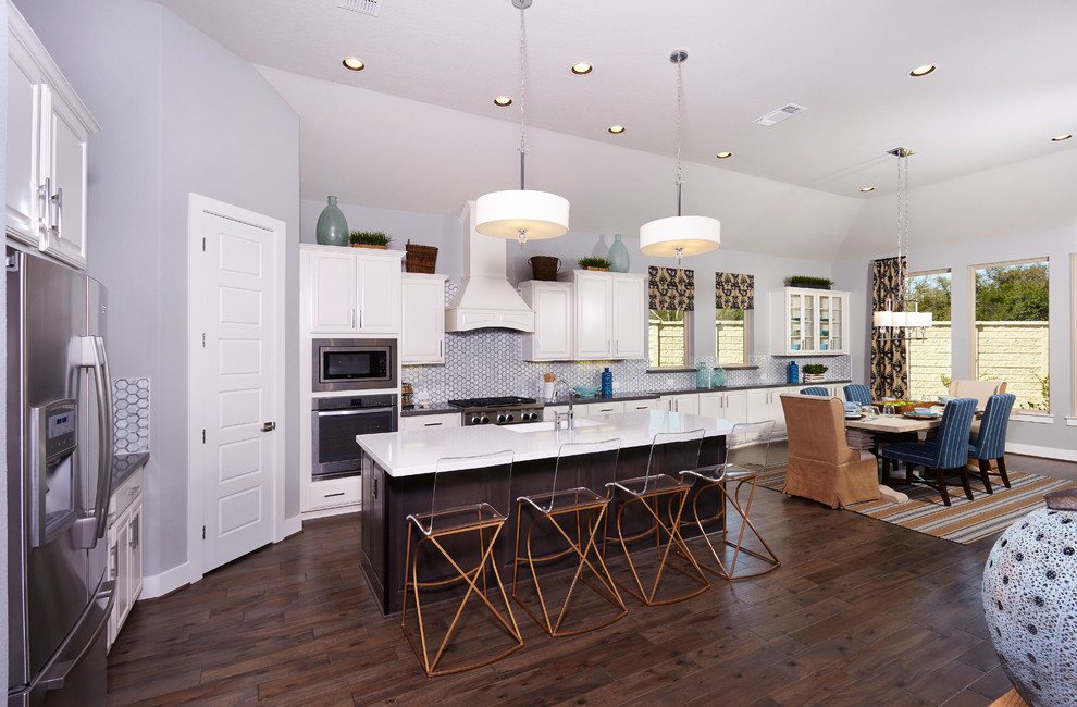 Large trendy l-shaped dark wood floor and brown floor eat-in kitchen photo in Austin with a farmhouse sink, raised-panel cabinets, white cabinets, solid surface countertops, gray backsplash, ceramic backsplash, stainless steel appliances and an island