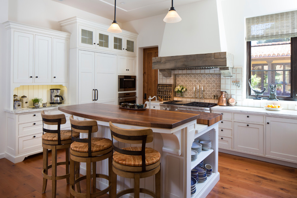 Mediterrane Küche mit Schrankfronten mit vertiefter Füllung, weißen Schränken, Küchenrückwand in Weiß, Küchengeräten aus Edelstahl und braunem Holzboden in Santa Barbara