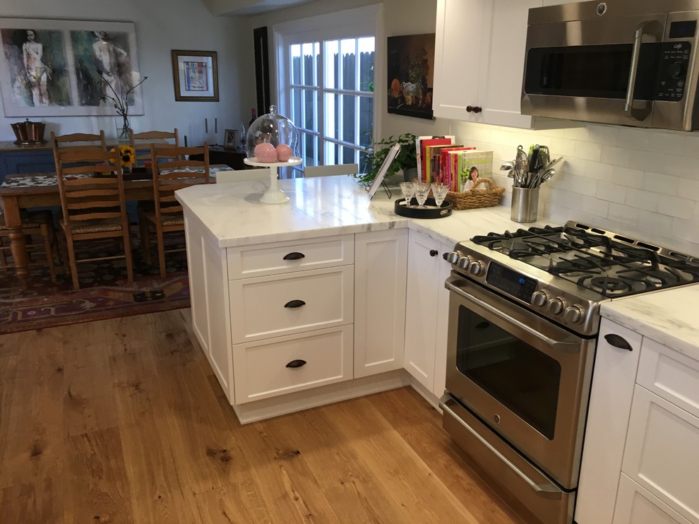 Large beach style l-shaped light wood floor eat-in kitchen photo in Orange County with a farmhouse sink, shaker cabinets, white cabinets, marble countertops, white backsplash, ceramic backsplash, stainless steel appliances and a peninsula
