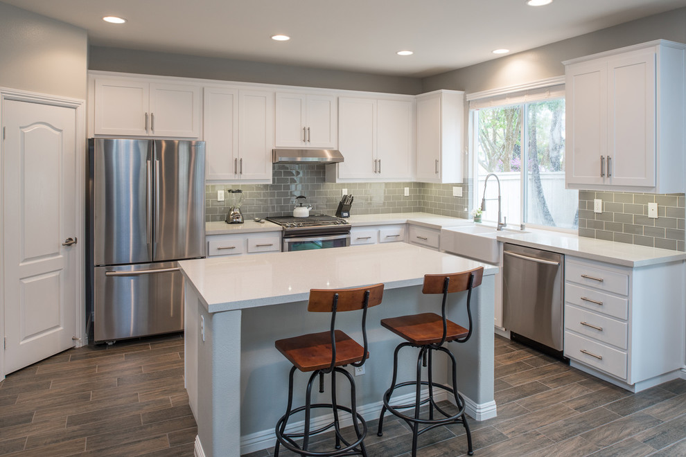 Example of a mid-sized minimalist u-shaped plywood floor kitchen design in San Diego with a farmhouse sink, shaker cabinets, white cabinets, quartz countertops, gray backsplash, subway tile backsplash, stainless steel appliances and an island