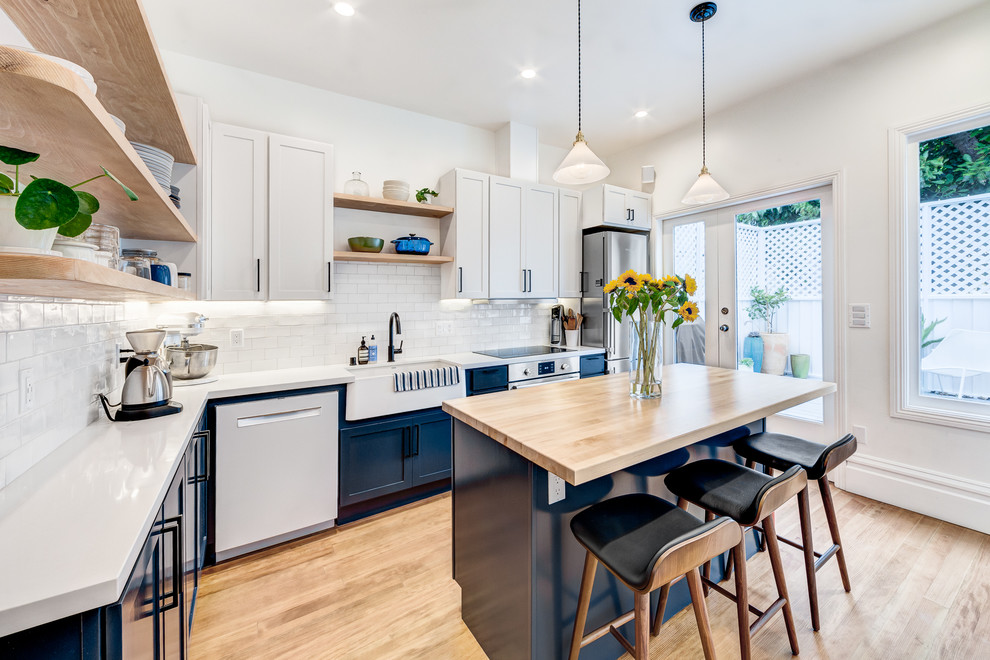 Idées déco pour une cuisine américaine campagne en L avec un évier de ferme, un placard à porte shaker, une crédence blanche, un électroménager en acier inoxydable, parquet clair, îlot, un sol beige et un plan de travail blanc.