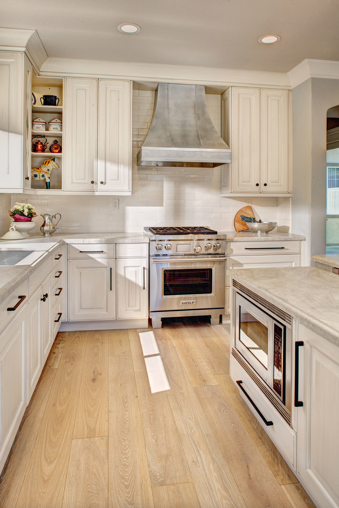 Mid-sized elegant l-shaped light wood floor open concept kitchen photo in Sacramento with an undermount sink, raised-panel cabinets, white cabinets, quartzite countertops, white backsplash, ceramic backsplash, stainless steel appliances and an island
