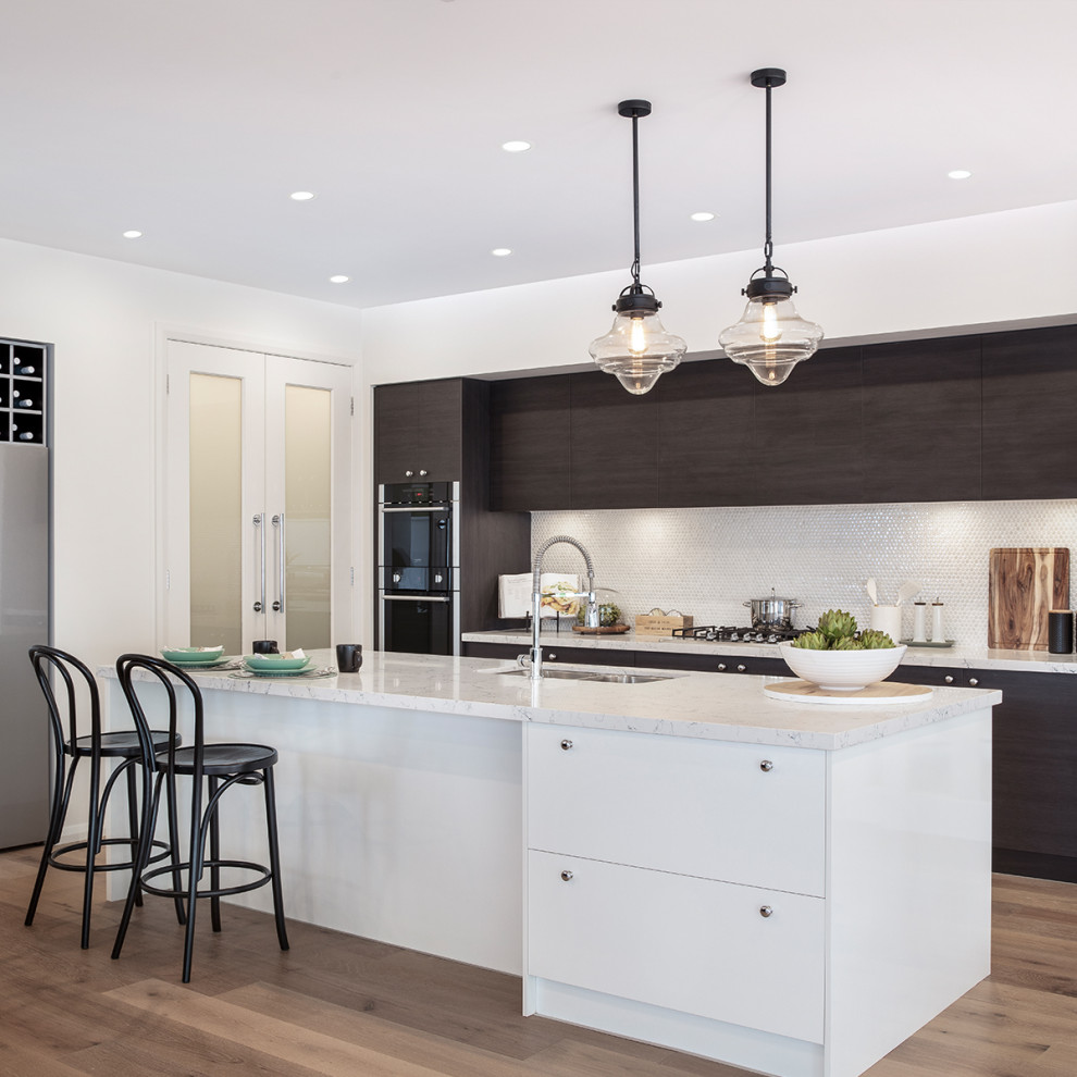 Design ideas for a large contemporary galley open plan kitchen in Sydney with brown cabinets, engineered stone countertops, white splashback, mosaic tiled splashback, medium hardwood flooring, an island, brown floors, white worktops, a submerged sink, flat-panel cabinets and black appliances.