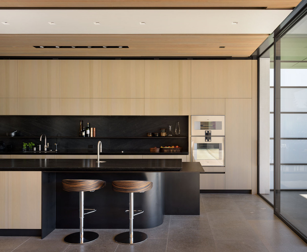 Cette image montre une cuisine parallèle minimaliste en bois clair avec un placard à porte plane, une crédence noire, un électroménager en acier inoxydable, îlot, un sol gris, plan de travail noir et un plafond en bois.