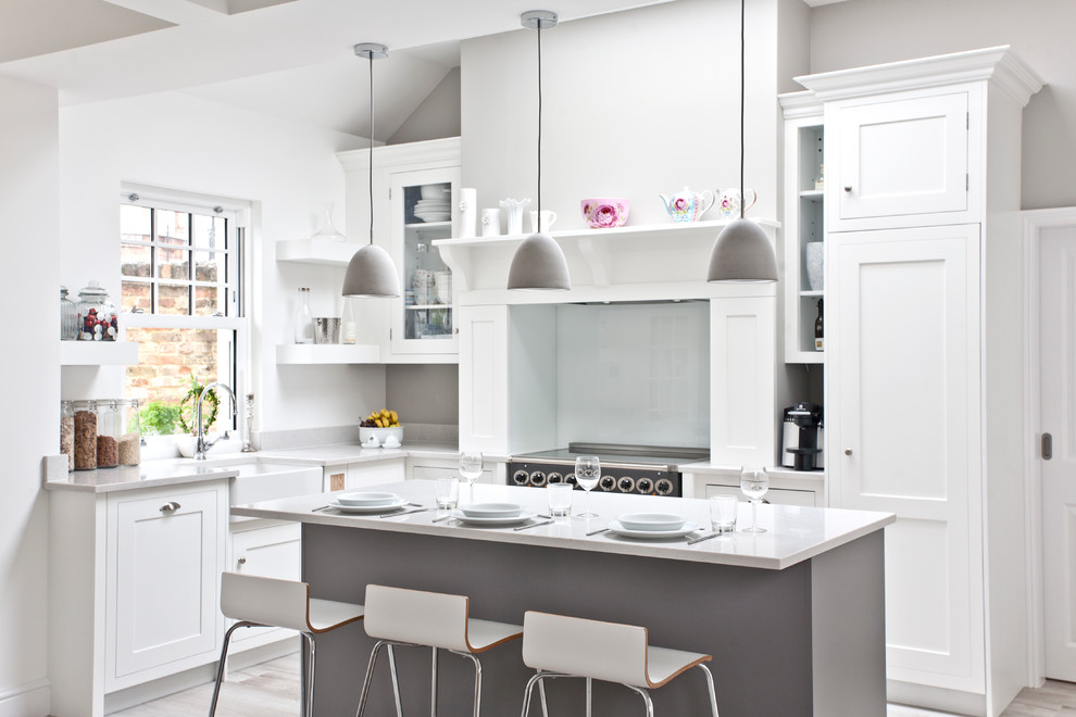 Example of a transitional l-shaped light wood floor eat-in kitchen design in Surrey with a farmhouse sink, an island, shaker cabinets, gray cabinets, solid surface countertops, gray backsplash, glass sheet backsplash and stainless steel appliances