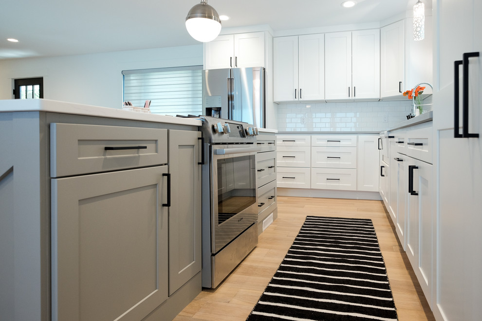 This is an example of a medium sized nautical l-shaped open plan kitchen in Grand Rapids with a belfast sink, shaker cabinets, white cabinets, engineered stone countertops, white splashback, metro tiled splashback, stainless steel appliances, light hardwood flooring and an island.